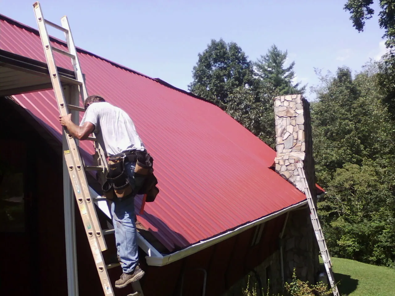Roofer on a ladder beside a red metal roof and stone chimney — Soffit & Fascia Repair by Roofing Brothers in Irvington